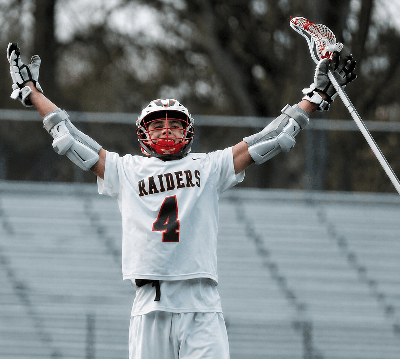 Charlie Griffith in a white Raiders lacrosse uniform, arms raised with his stick after a goal.