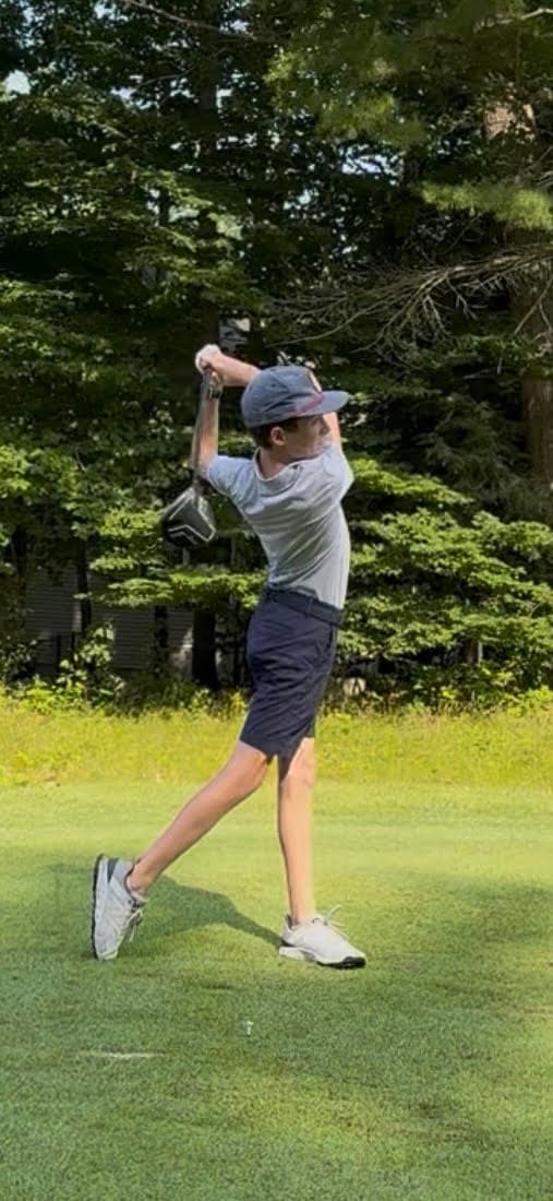 Gage Olcott at the top of his follow-through after a tee shot on a tree-lined golf course.