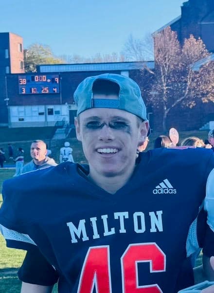 Taylor Andrews smiling on the sideline in a navy Milton football jersey and a backwards cap.