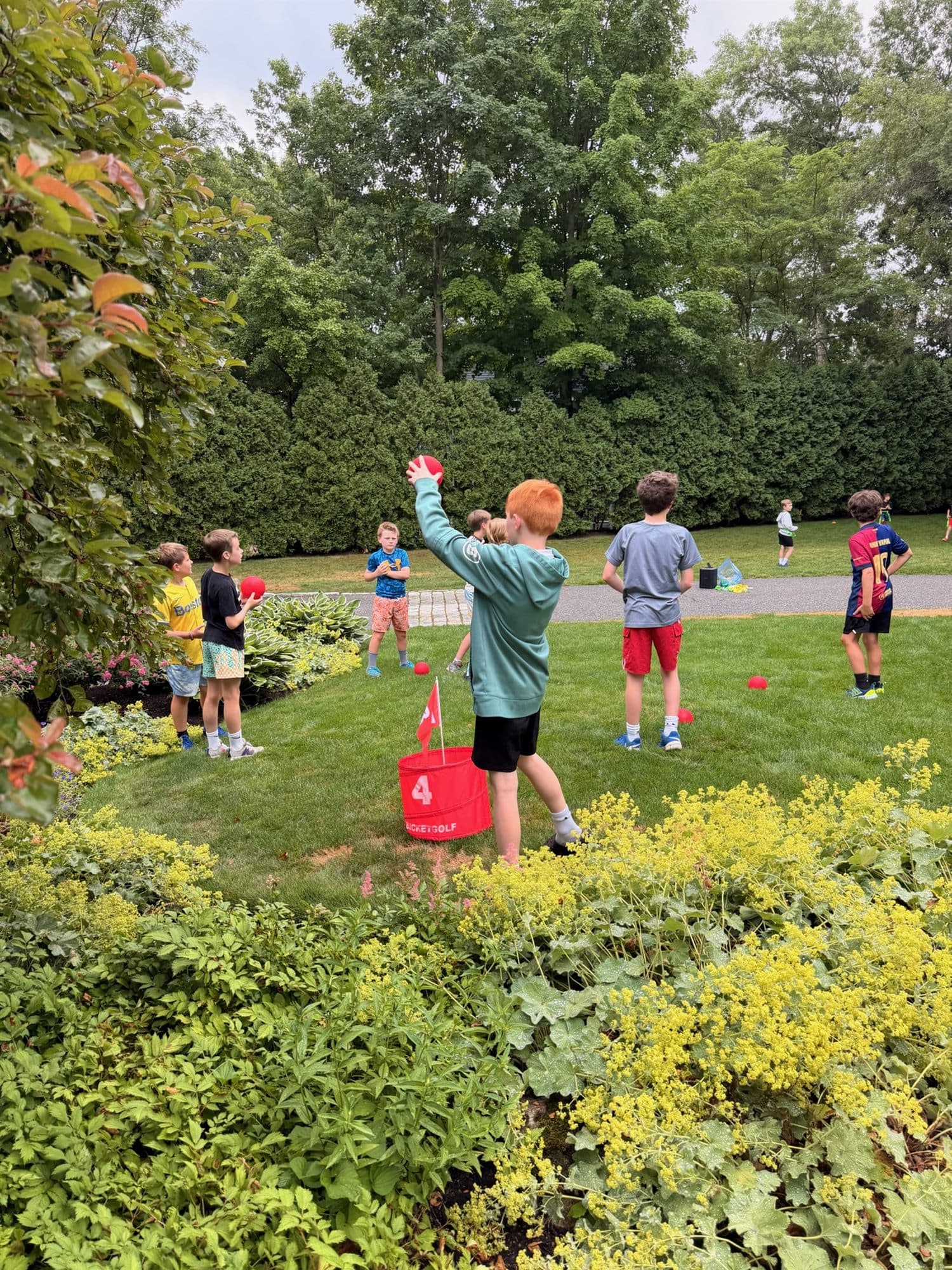 A camper winding up to throw at a red target bucket marked 4 on the lawn while other campers wait their turn.
